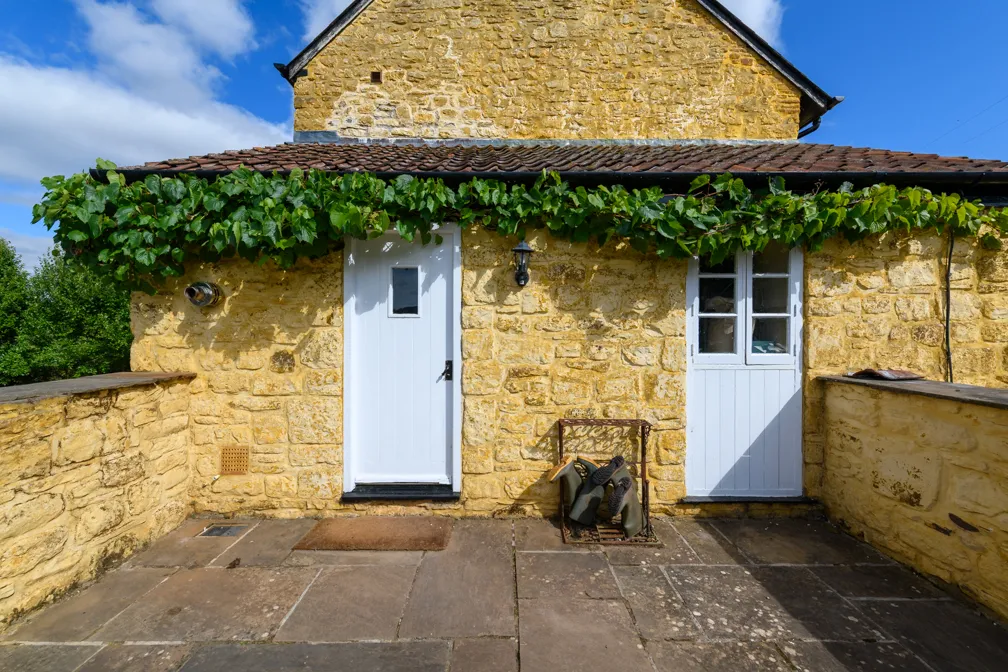 Stone house with two white doors, a green vine growing above them, and a metal rack holding garden boots on a paved patio.