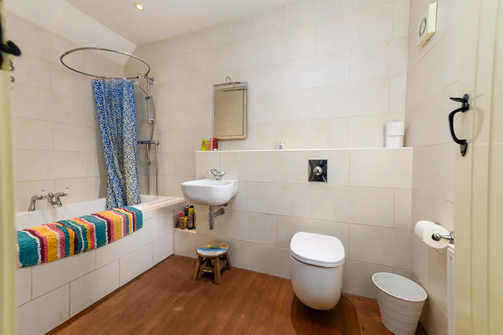 Modern bathroom with beige tiled walls, wooden floor, bathtub with colorful striped towel, wall-mounted sink, small wooden stool, and a white toilet.