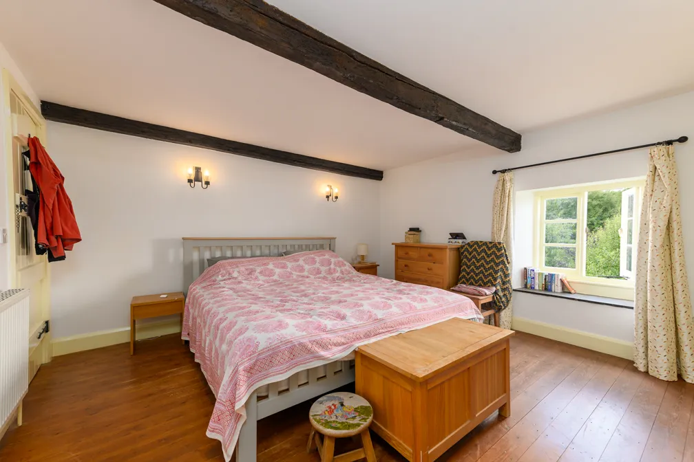 Cozy bedroom with a large bed covered with a pink floral bedspread, wooden chest at the foot, and a window with floral curtains letting in natural light.
