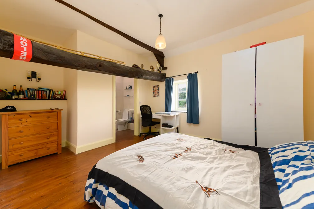 Bedroom with wooden dresser, desk and chair by window, white wardrobe, bed with a patterned duvet, and visible bathroom through open door.