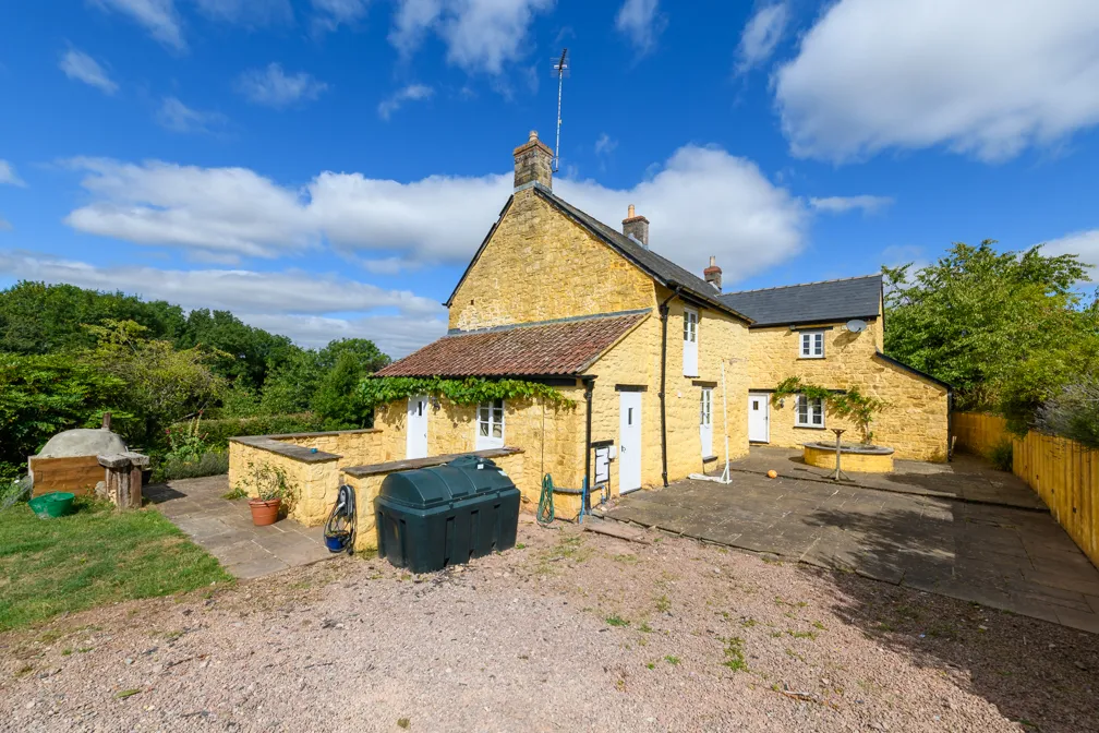 Stone country house with yellow walls, white doors, and a tiled roof under a blue sky with scattered clouds.