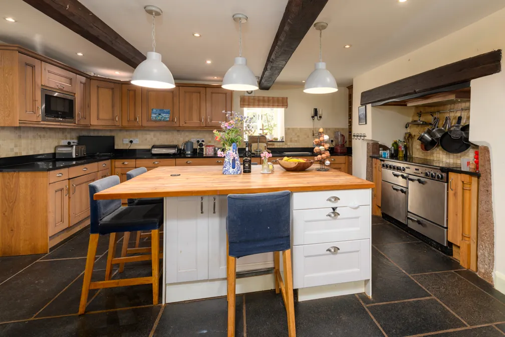 Cozy kitchen with wooden cabinets, black countertops, a large wooden island with blue cushioned stools, and hanging pots by the stove.
