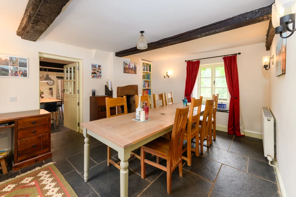 Cozy dining room with a wooden table and eight chairs, exposed dark wooden ceiling beams, red curtains on window, and a door leading to the kitchen.