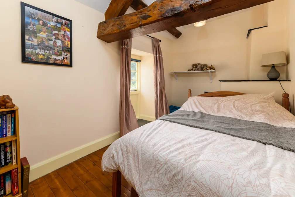Cozy bedroom with wooden beams, a bed with floral-patterned bedding, grey throw, lamp on corner shelf, and framed photo collage on wall.