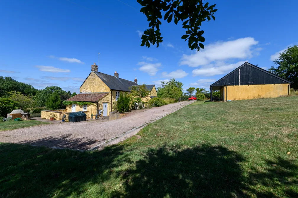 Yellow stone house with a gravel driveway, adjacent grassy field, an outbuilding, and a clear blue sky with scattered clouds.