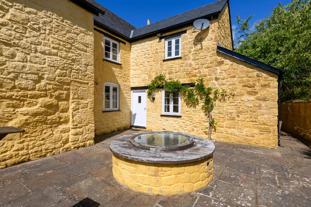 Stone courtyard with a round covered well in front of a yellow stone house with white framed windows and a small climbing plant on the wall.