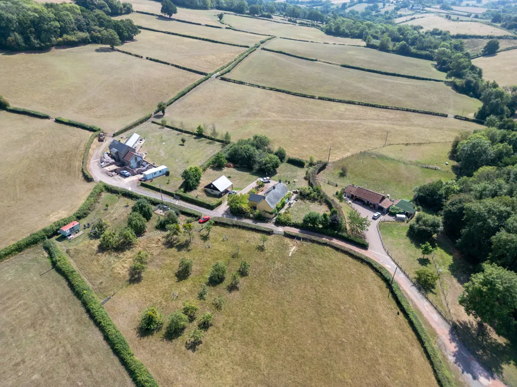 Aerial view of a rural countryside with fields divided by hedgerows, featuring a few houses, trees, and parked cars along narrow roads.
