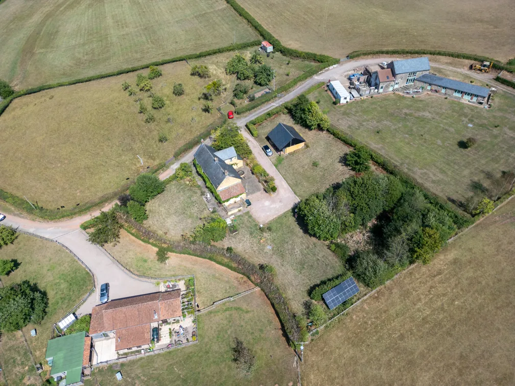 Aerial view of rural houses with gardens, trees, solar panels, and surrounding farmland separated by hedges.