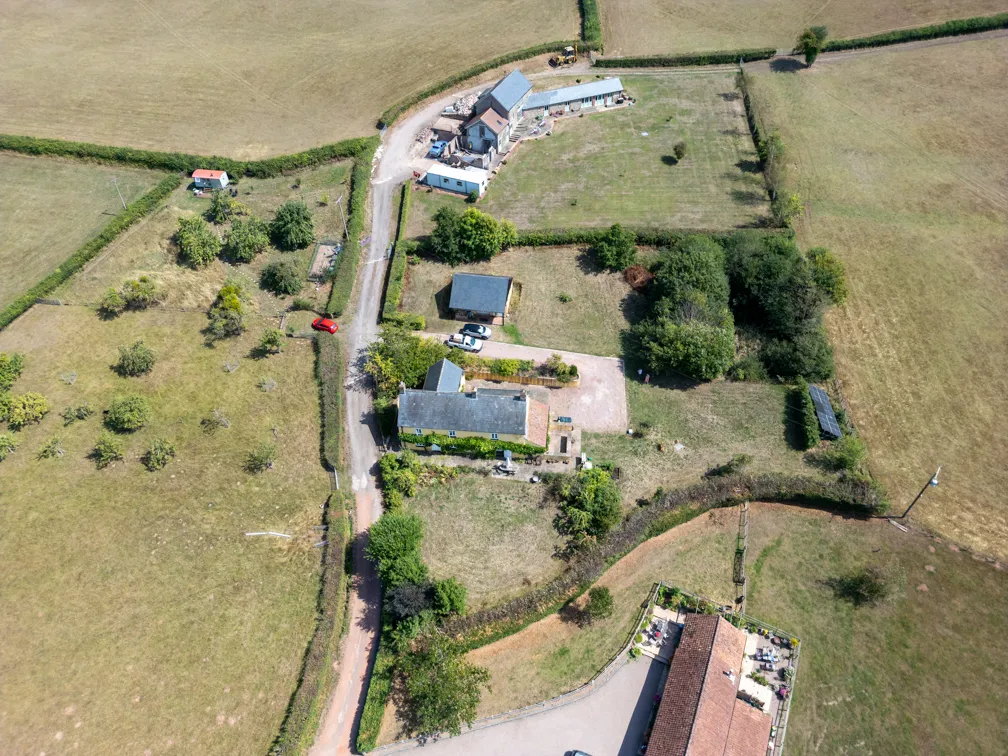 Aerial view of rural property with several buildings, a driveway, parked cars, and surrounding green fields divided by hedges.
