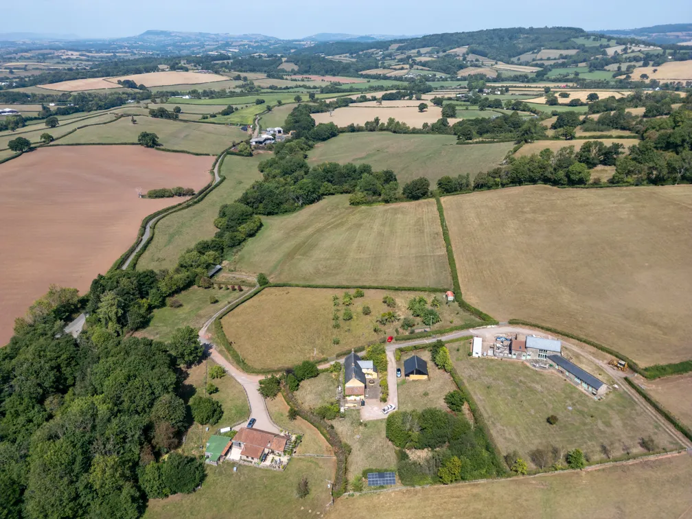 Aerial view of rural farmland with scattered houses, fields bordered by hedges, and rolling hills in the background.