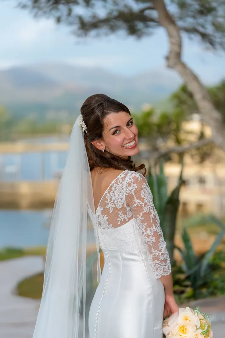 Smiling bride in a white lace wedding dress with a long veil holding a bouquet of yellow and white roses outdoors.