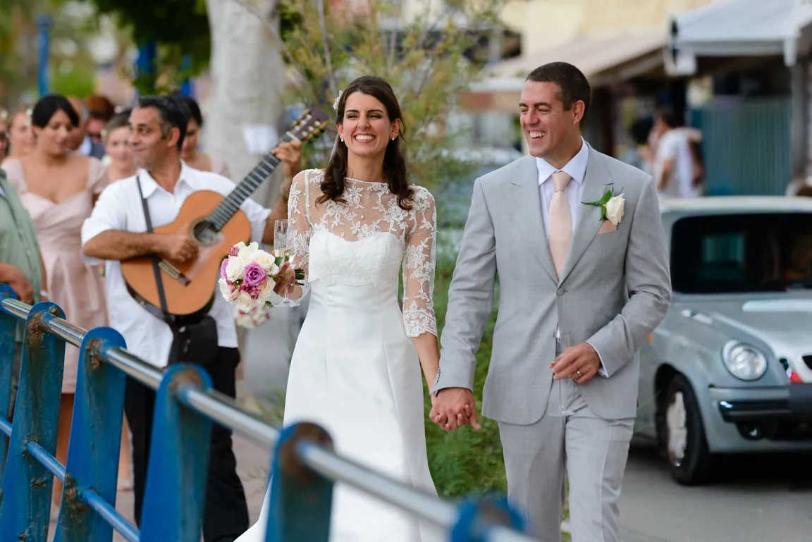 Bride in white dress and groom in light gray suit holding hands and smiling while walking outdoors with a guitar player behind them.
