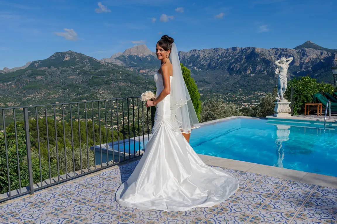 Bride in a white wedding dress holding a bouquet standing on a tiled patio next to a pool with mountains in the background.