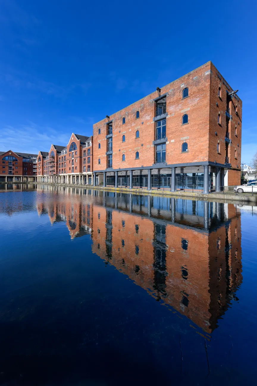 Red brick buildings with arched windows reflecting in calm water under a clear blue sky.