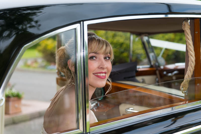Smiling bride with blonde hair sitting inside a vintage car, seen through the passenger window.
