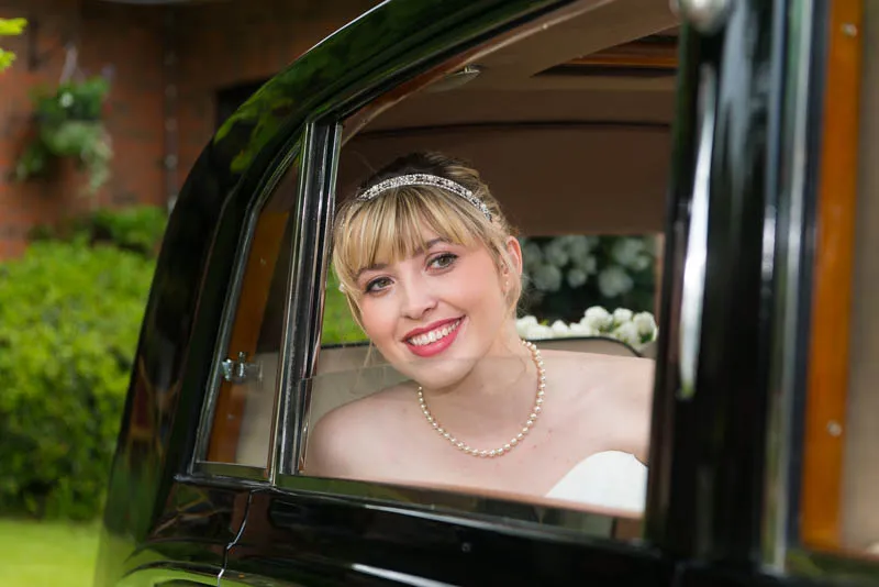 Smiling bride wearing a pearl necklace and tiara, looking out from a vintage car window.