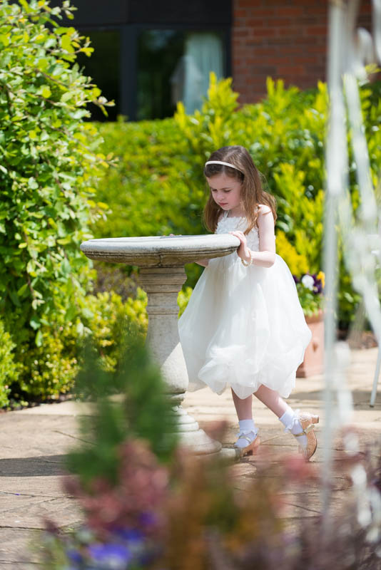 Young girl in a white dress and headband leaning on a stone birdbath in a sunlit garden.