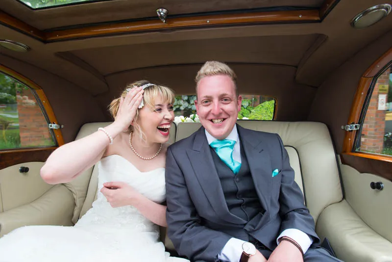 Smiling bride in a white wedding dress and groom in a gray suit with turquoise tie sitting together in the backseat of a car.