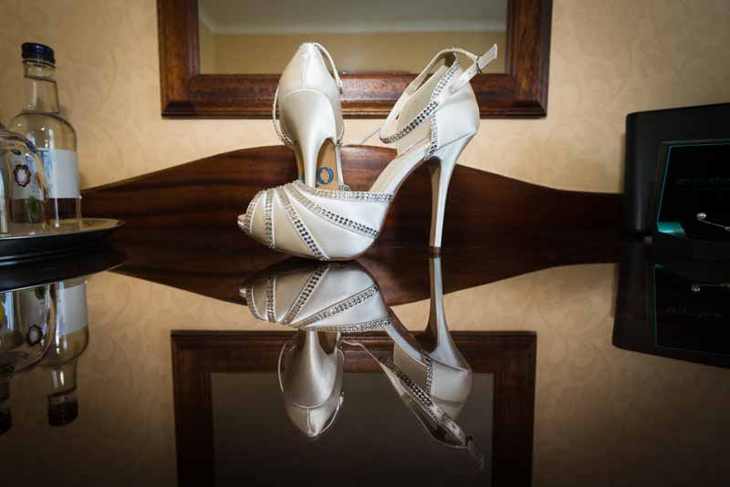 Pair of elegant white high-heeled shoes with rhinestone straps reflected on a polished dark surface.