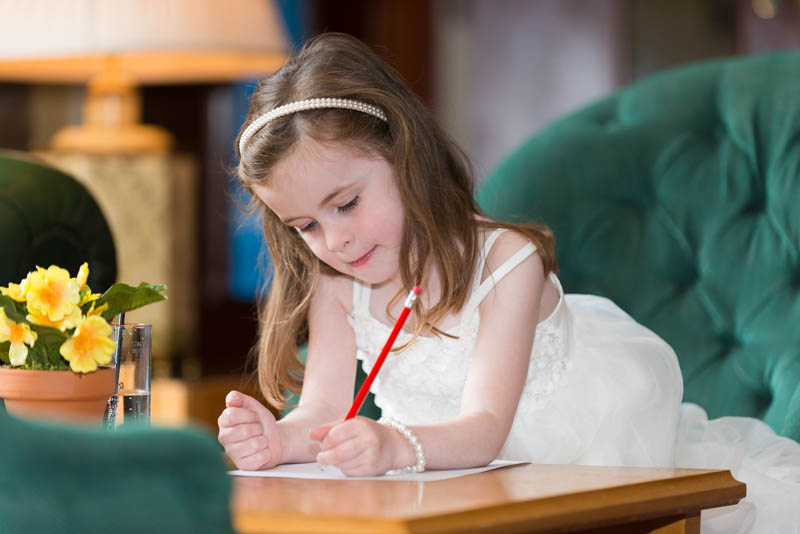 Young girl in a white dress with a pearl headband and bracelet, leaning on a table while drawing with a red pencil in a cozy living room.