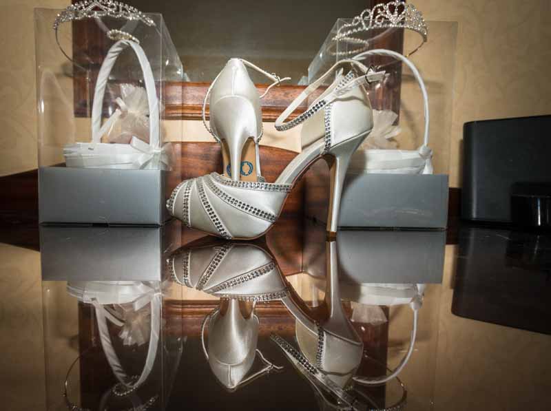 Pair of white high-heeled wedding shoes with rhinestone accents reflected on a glossy surface, flanked by two clear boxes containing white headbands with tiaras.