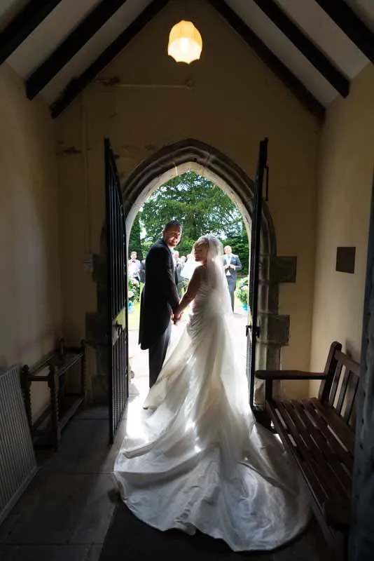 Bride and groom holding hands and smiling while standing at an open church doorway with bright outdoor light and greenery behind them.