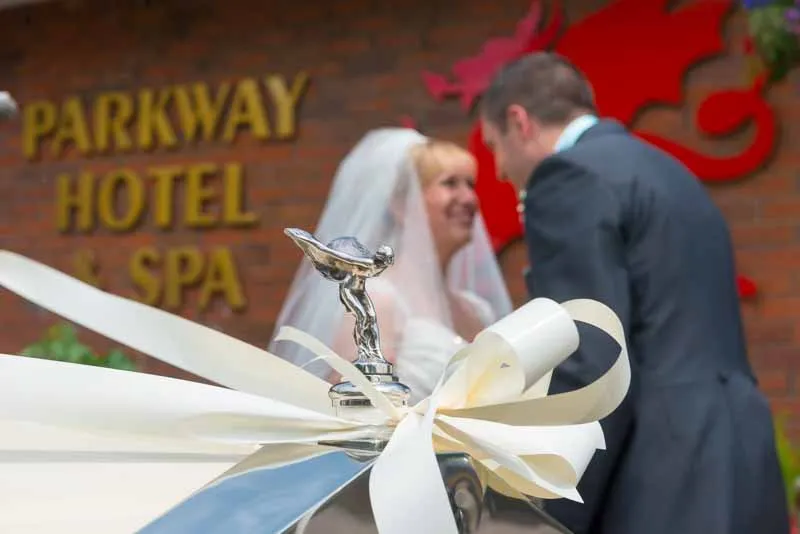 Close-up of a Rolls-Royce hood ornament with a white ribbon, a bride and groom smiling in the blurred background outside Parkway Hotel & Spa.