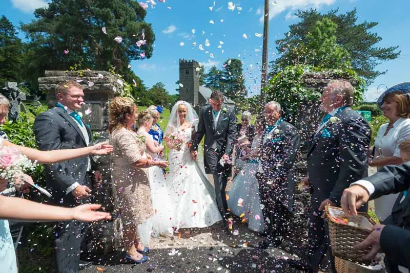 Bride and groom walking hand in hand outdoors while guests throw flower petals in celebration.