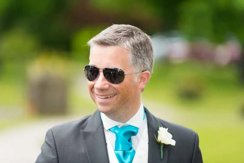 Man wearing a dark suit, bright turquoise tie, and sunglasses smiling outdoors with a blurred green background.