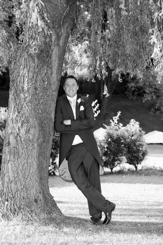 Man in formal attire leaning against a large tree in a park, smiling and with arms crossed.