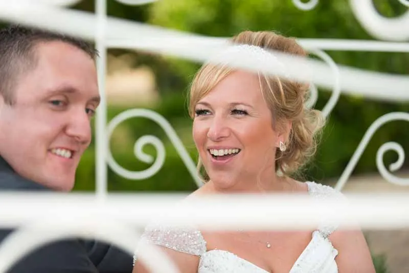 Smiling bride in a white dress with a groom, framed by white decorative metal bars.