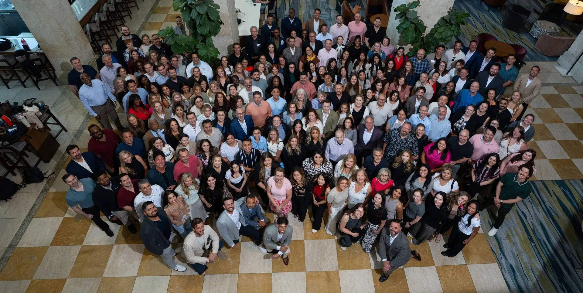Large group of diverse people standing together indoors on a checkered tile floor, looking up at the camera.