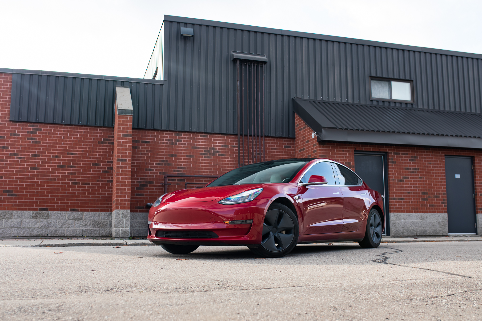 Red Tesla Model 3 parked in front of a red brick building with black siding. The car is protected by ExoShield Windshield Protection Film