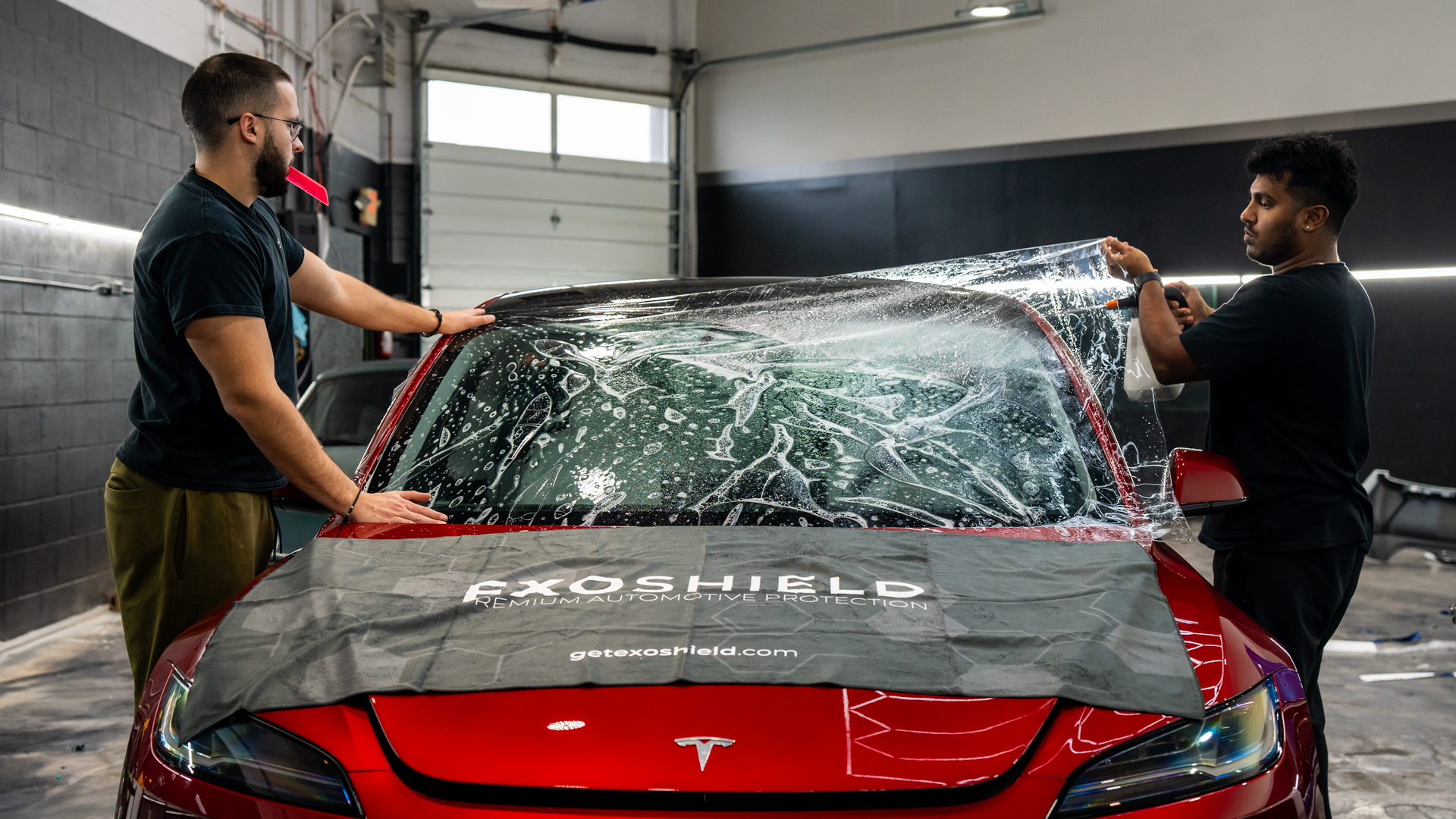 Red Tesla M3 receiving SPRINT WPF from ExoShield from two people on either side of the car in a well-lit auto-body shop. 