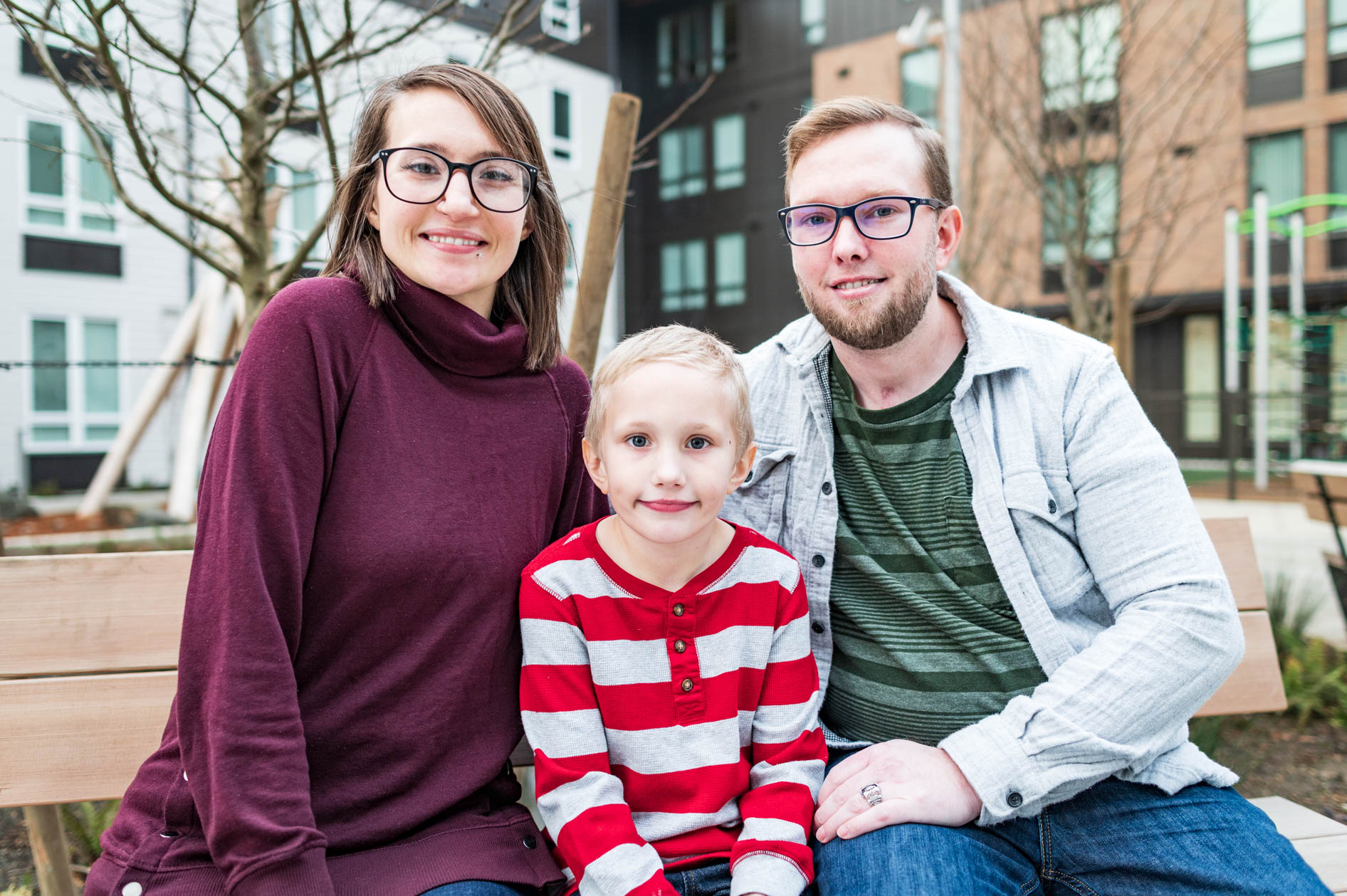 A portrait of a family of three—two adults and a child—sitting on a bench in a modern urban residential area. They are all wearing glasses and smiling, representing a narrative of stable, modern family life.