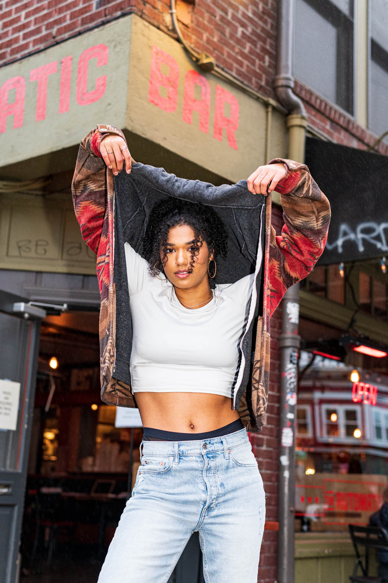 Young woman with curly hair lifting her jacket hood standing outside a brick bar with neon open sign.