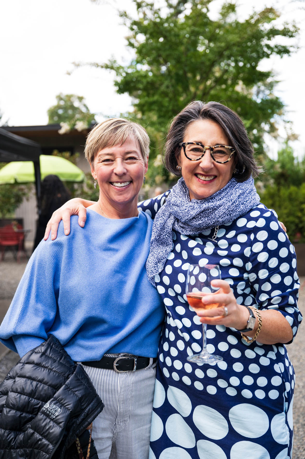 Two smiling women posing outdoors, one holding a wine glass and the other holding a jacket.