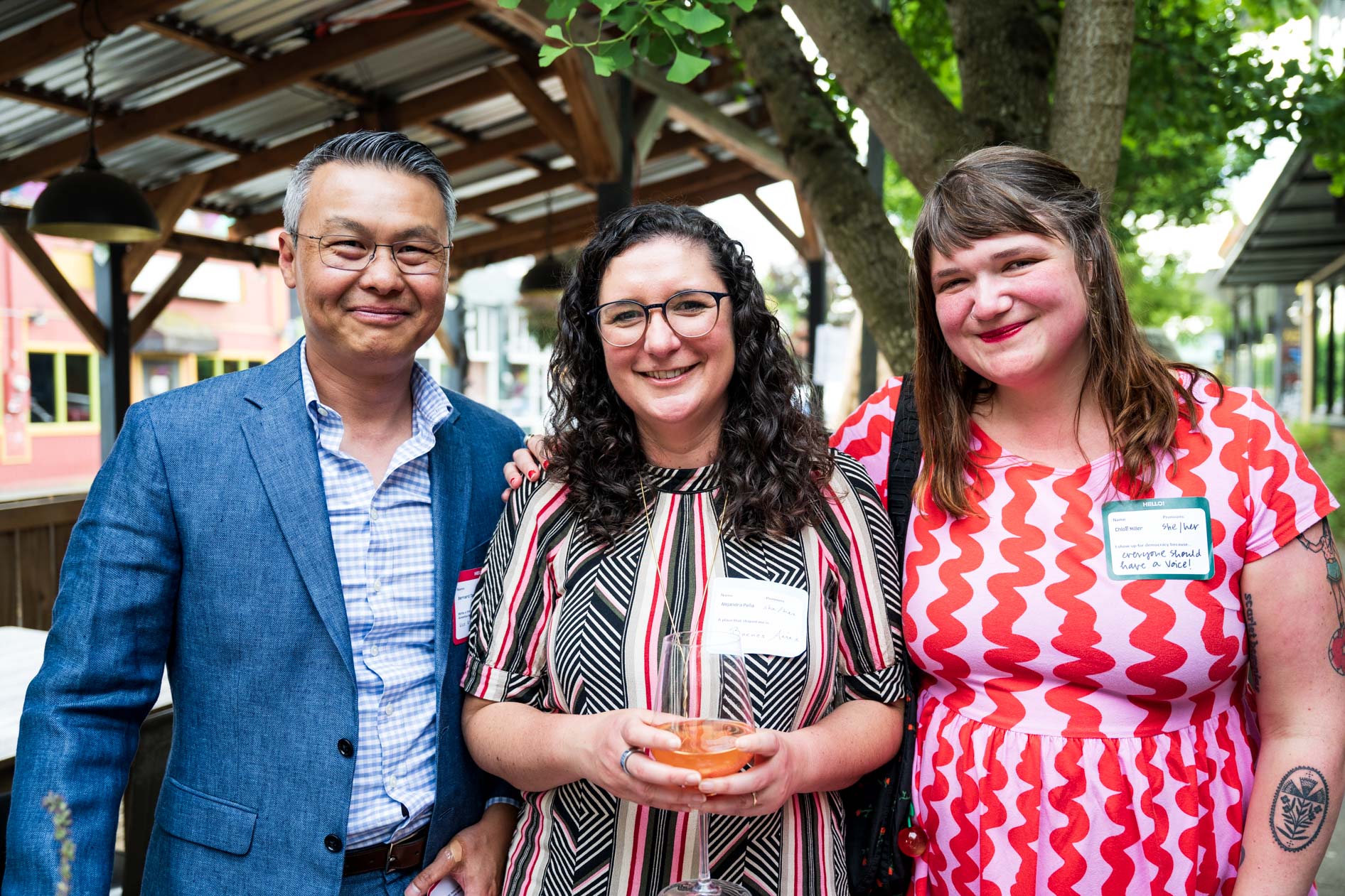 Three people smiling outdoors under a wooden canopy, one holding a drink, wearing name tags.