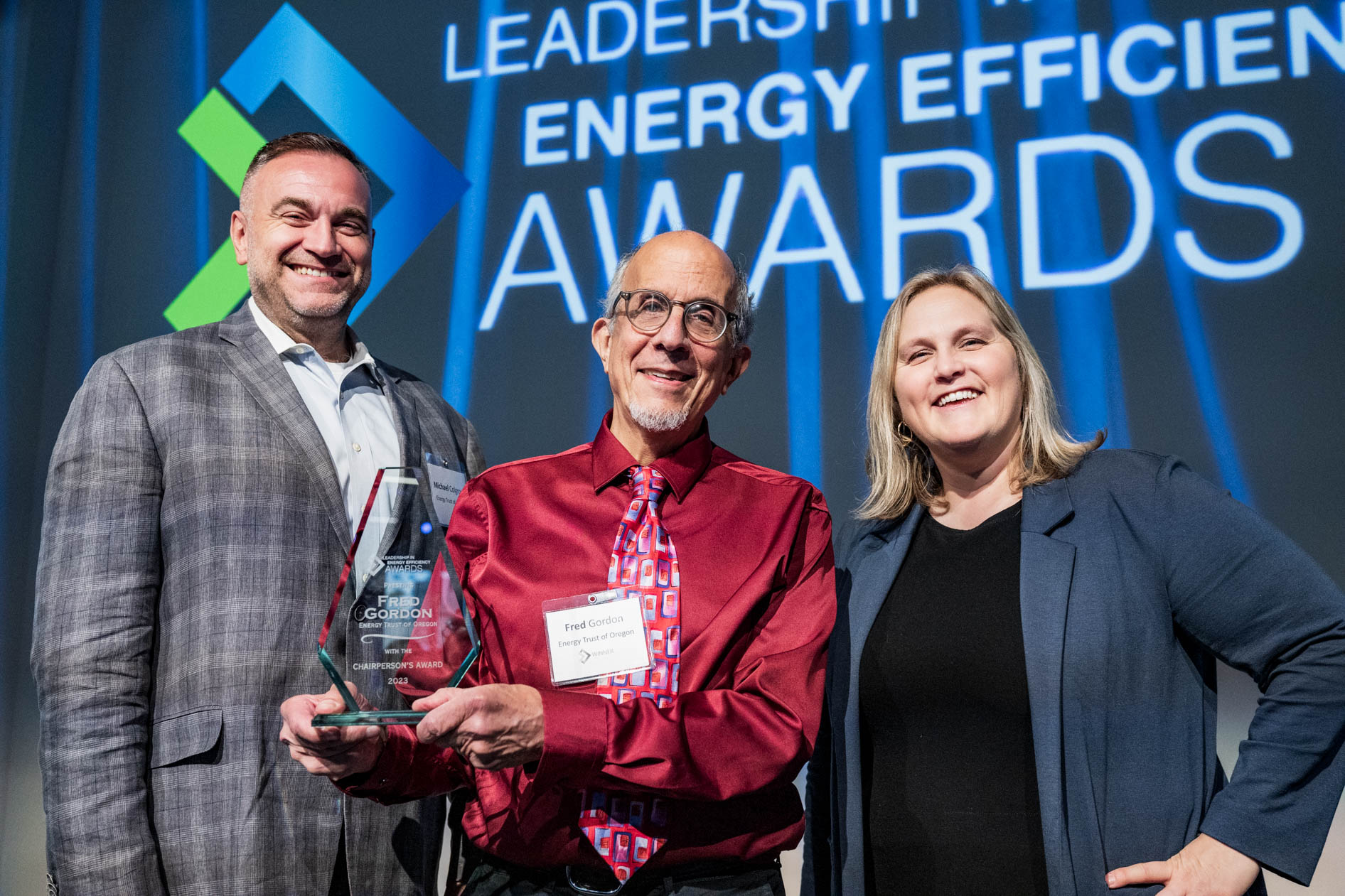 Three people smiling at an awards event; man in center holds a glass award trophy.