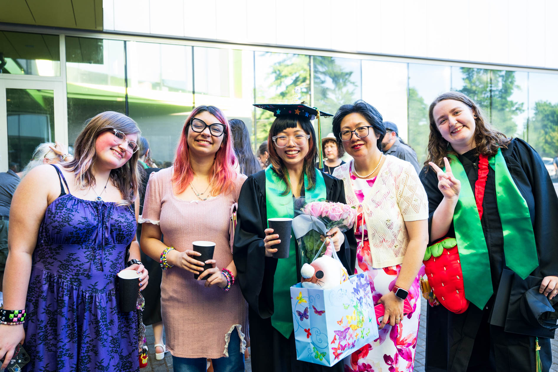 Group of five smiling women, two in graduation gowns holding flowers and gifts, outside building.
