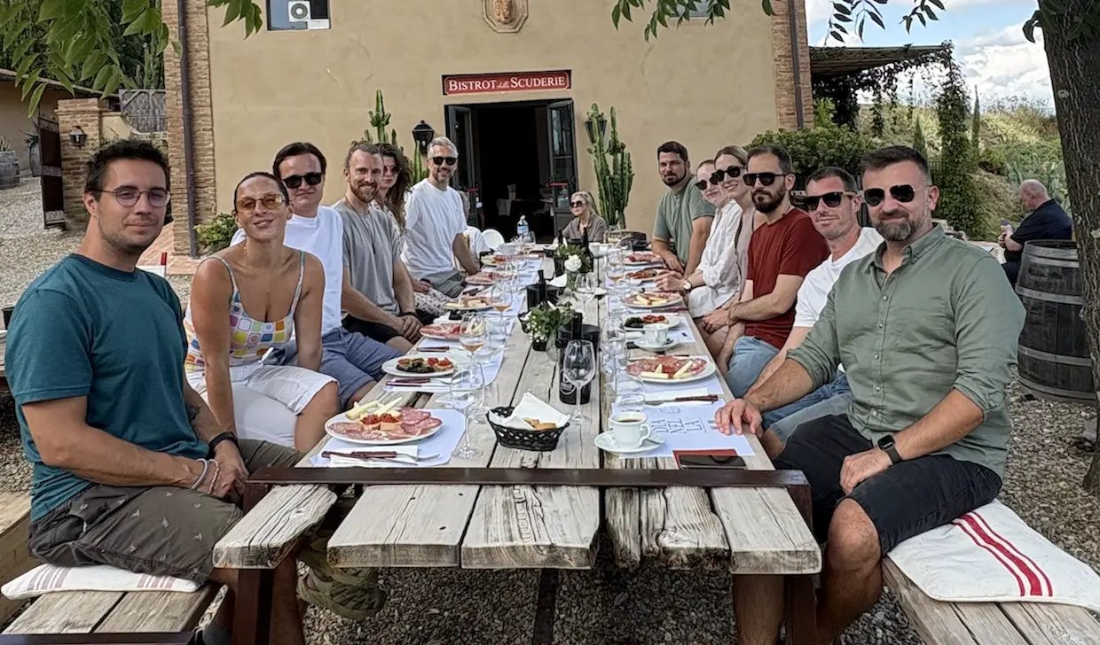 Group of people smiling while seated around a long outdoor wooden table with plates of food in front of a rustic building.