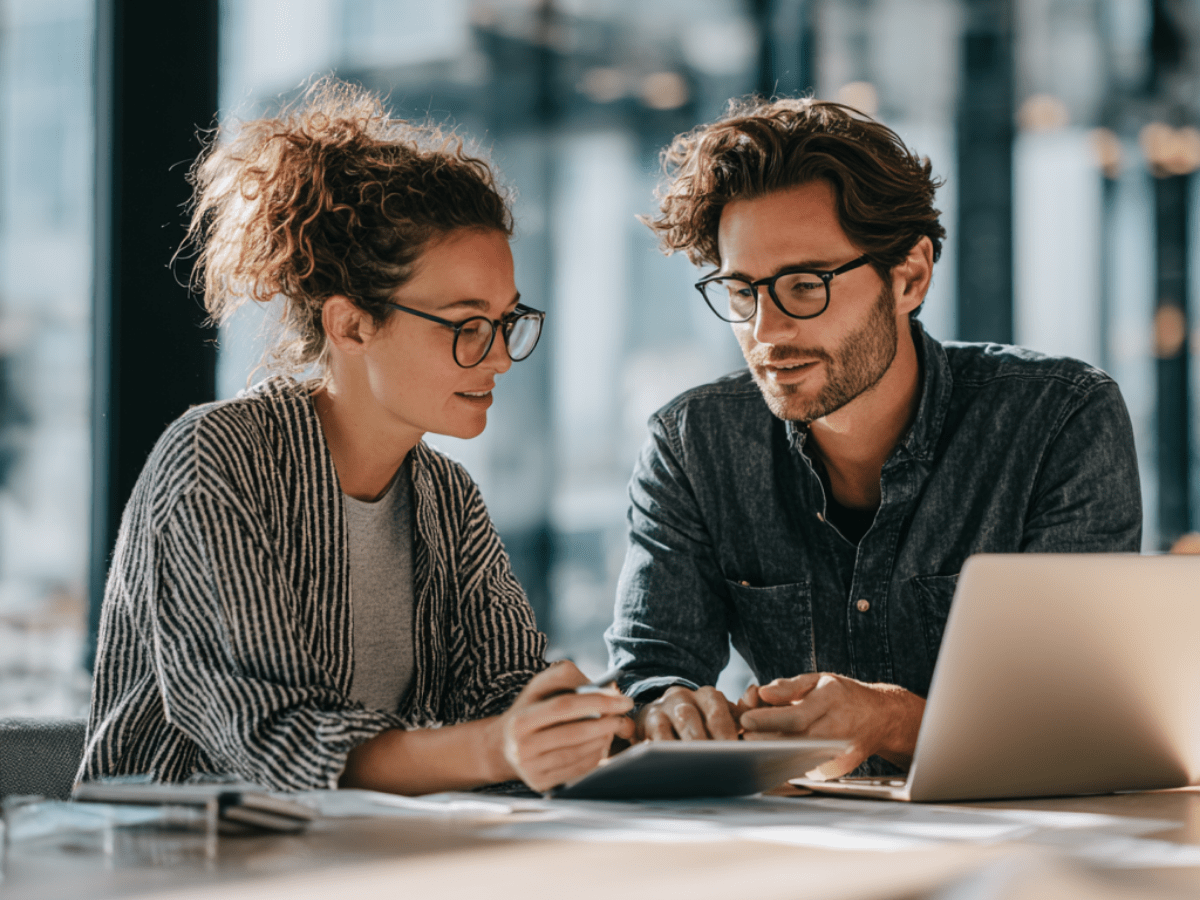 Two colleagues wearing glasses collaborating at a desk with a tablet and laptop, discussing project strategy and planning in a bright, modern office environment.