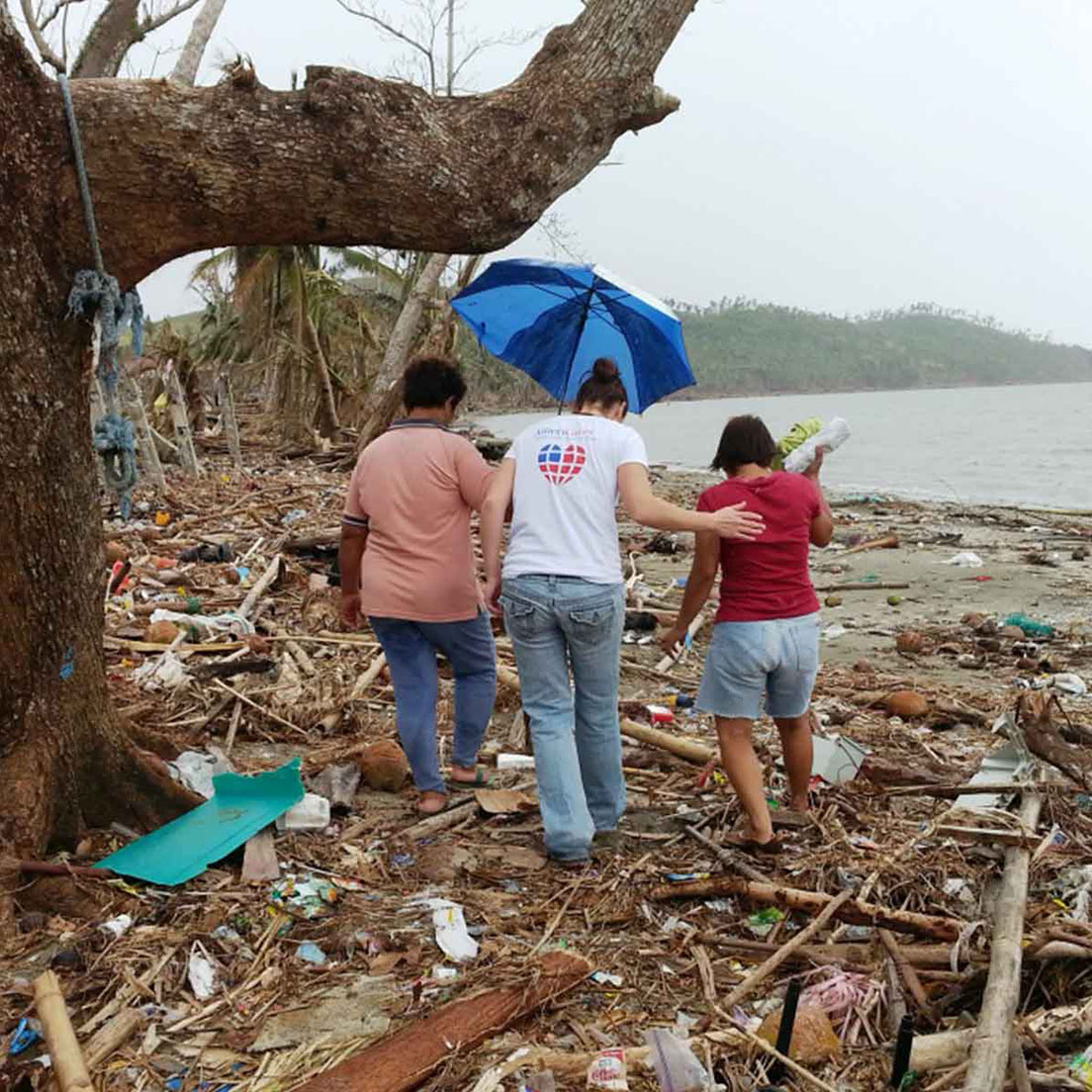 Three people walk together along a debris-covered coastline after a storm, arms linked for support. The person in the center wears an Americares T-shirt and holds a blue umbrella overhead, while the woman on the left in a pink shirt and the woman on the right in a red shirt walk alongside her. The beach is littered with broken branches, uprooted trees, and storm wreckage, with a gray sea and distant hills visible in the background.