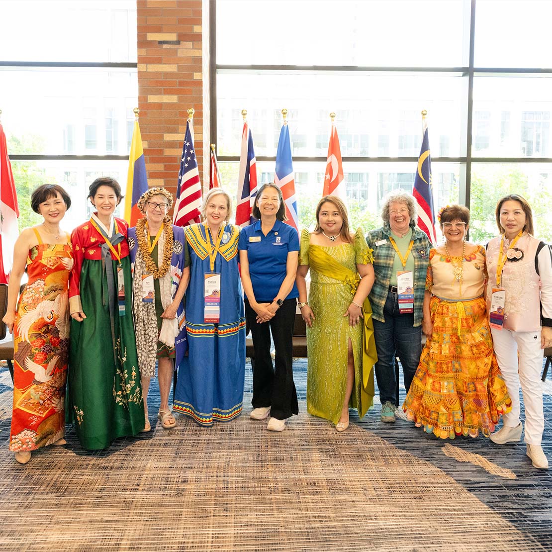 A diverse group of women in traditional dress from around the world stand together smiling in front of a row of international flags at a Soroptimist event.