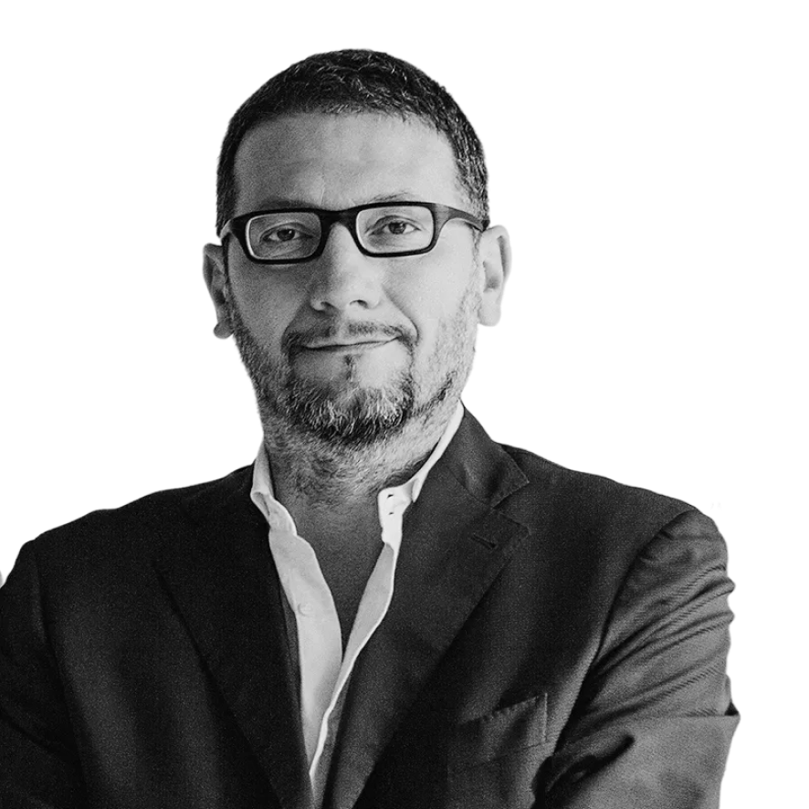 Man with short hair and beard wearing glasses, a white shirt, and a dark blazer, smiling slightly with arms crossed.