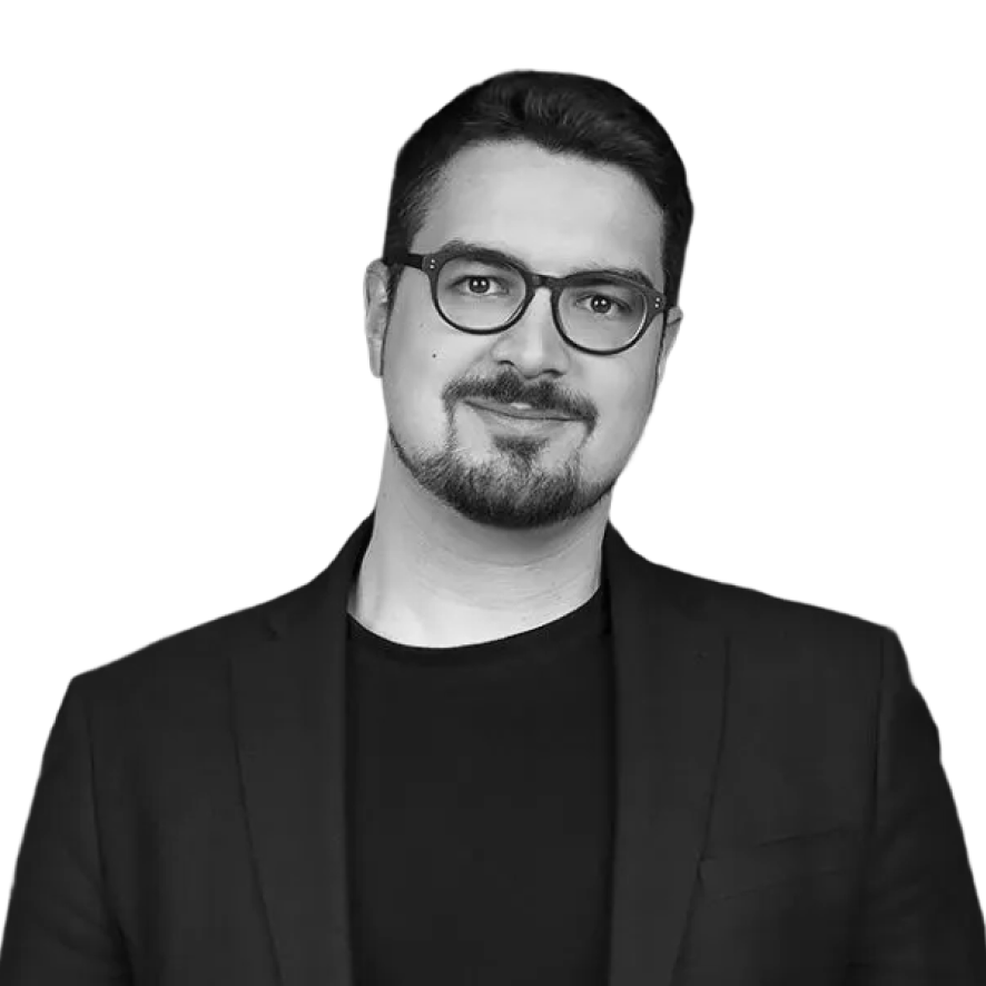 Portrait of a man with glasses, short dark hair, and a beard, wearing a black blazer and shirt, smiling slightly.