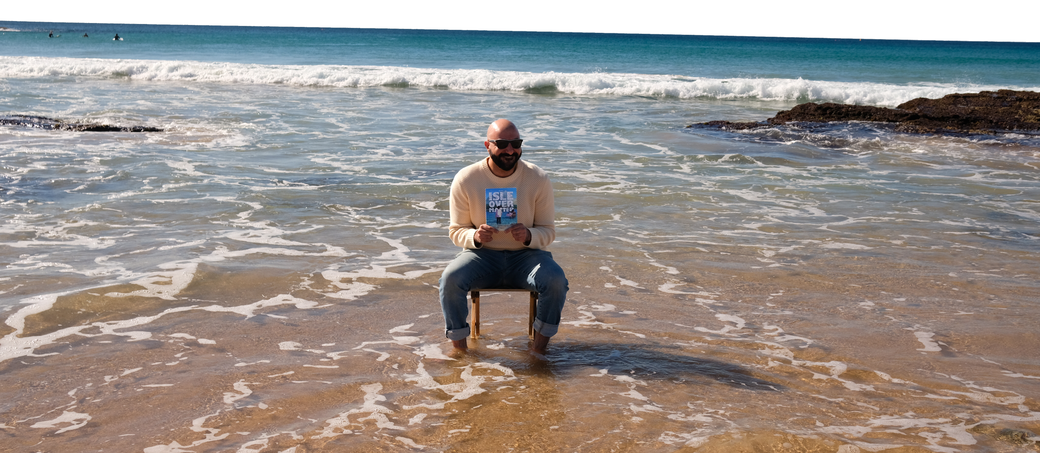 A man wearing sunglasses and a beige sweater sits on a small wooden stool in shallow seawater at the beach, holding a book titled 'ISLE OVER MATTER' with waves and rocks in the background.