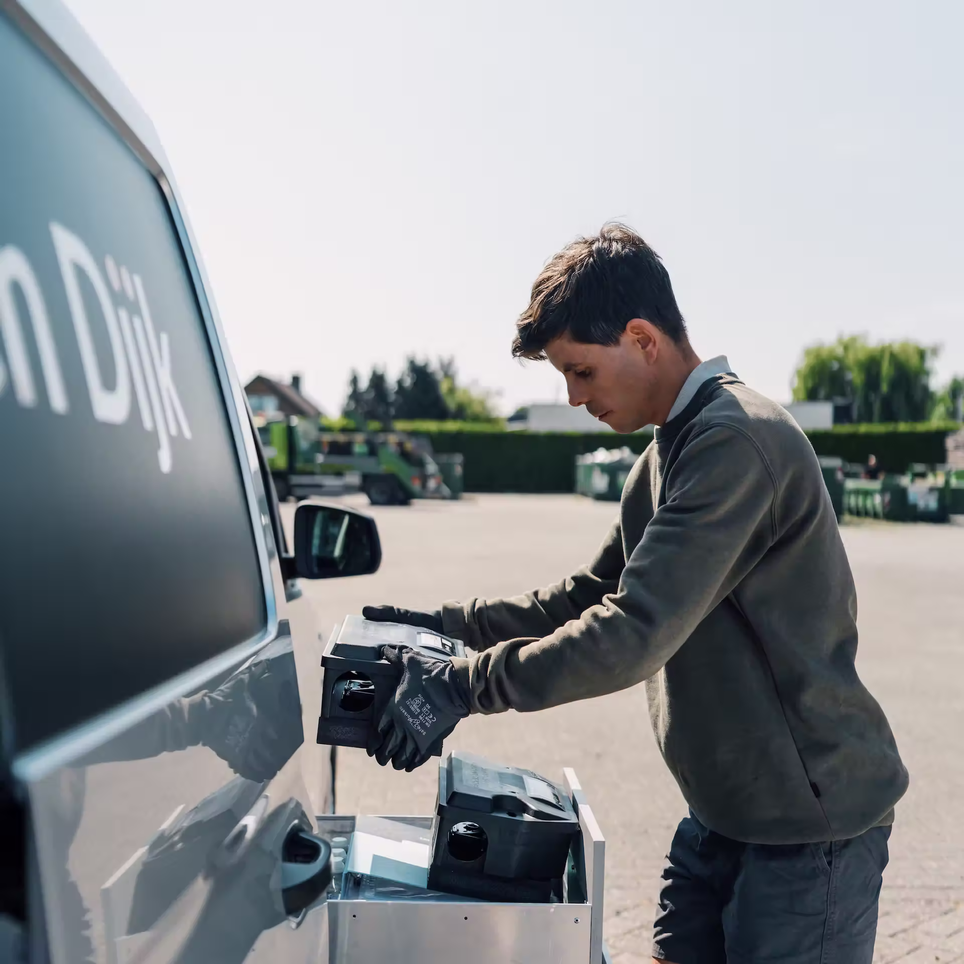 Jonge man met handschoenen plaatst apparaten in laadruimte van bedrijfswagen op een zonnige dag.