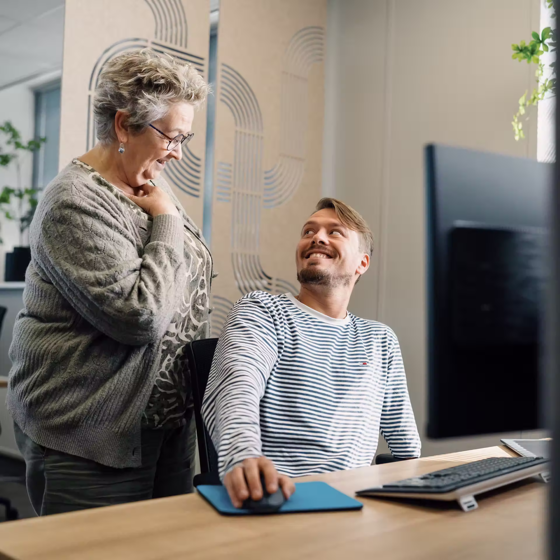 Een oudere vrouw glimlacht naar een jongere man die aan een bureau met een computer zit en teruglacht.