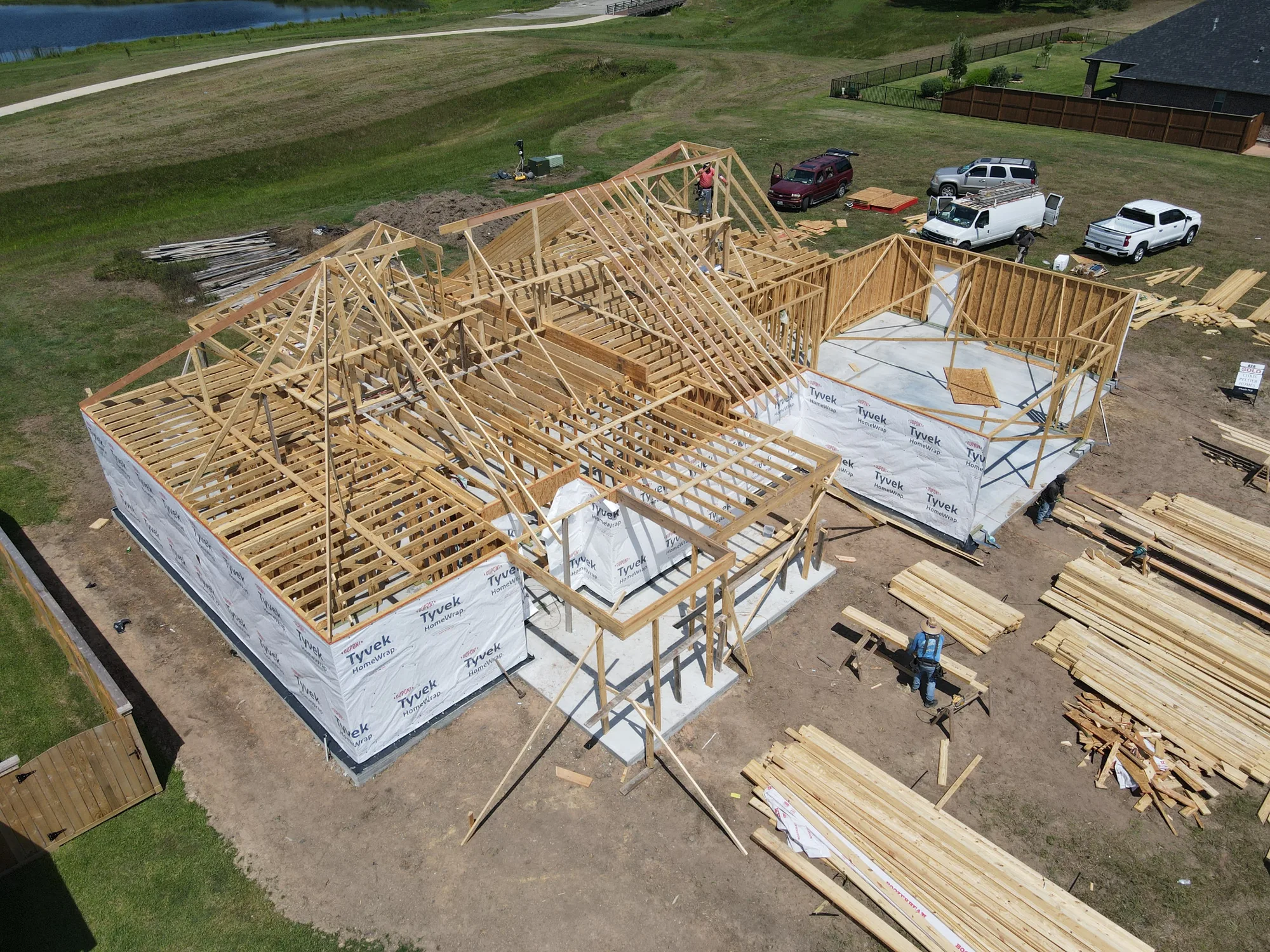 Aerial view of a house under construction showing wooden framing for walls and roof over a concrete foundation, with workers on site, stacks of lumber, and several parked vehicles nearby on a grassy plot.